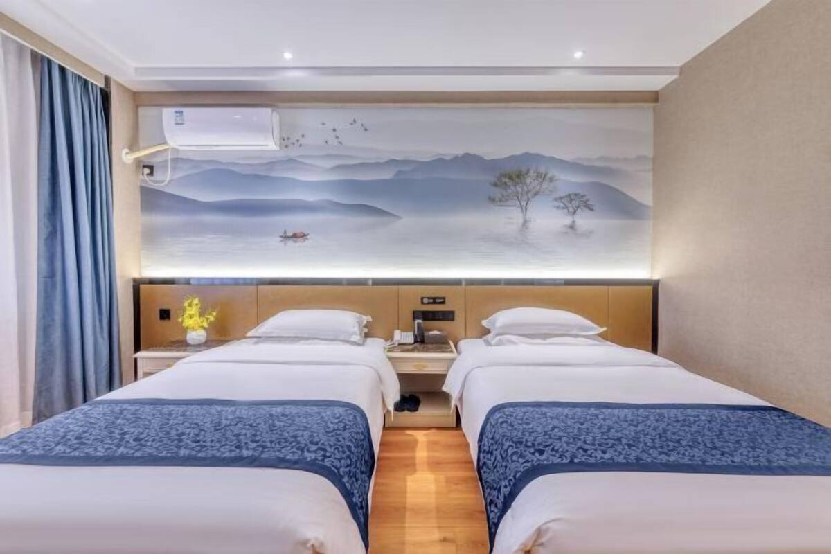 A hotel room with two beds, a mural on the wall, a bedside table with a lamp, and an air conditioning unit.