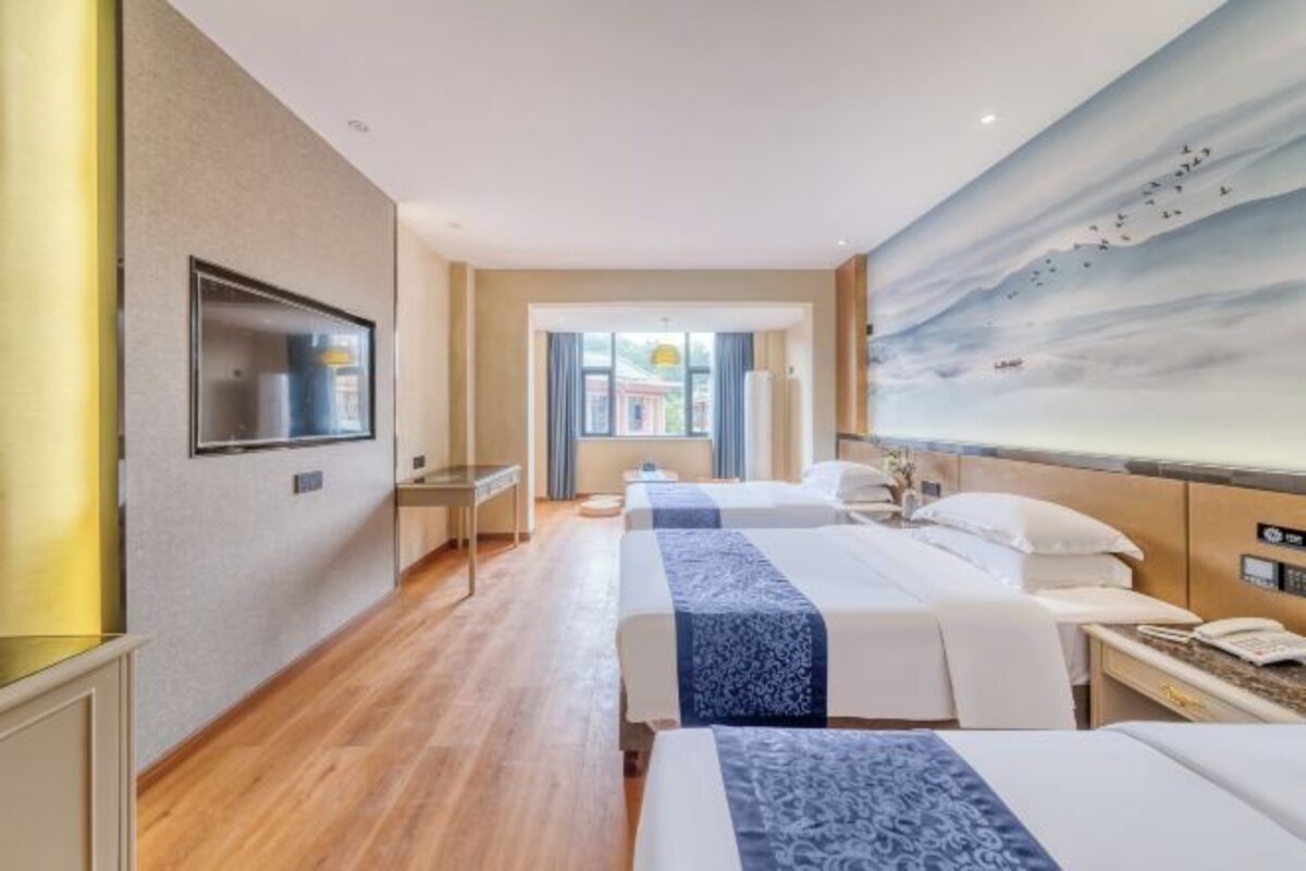 A hotel room with two beds, a TV mounted on the wall, a painting of birds, and a view through the window.