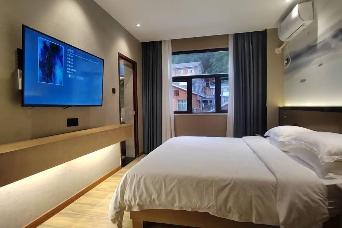 A hotel room with a bed, a TV mounted on the wall, a window with curtains, and an air conditioning unit.