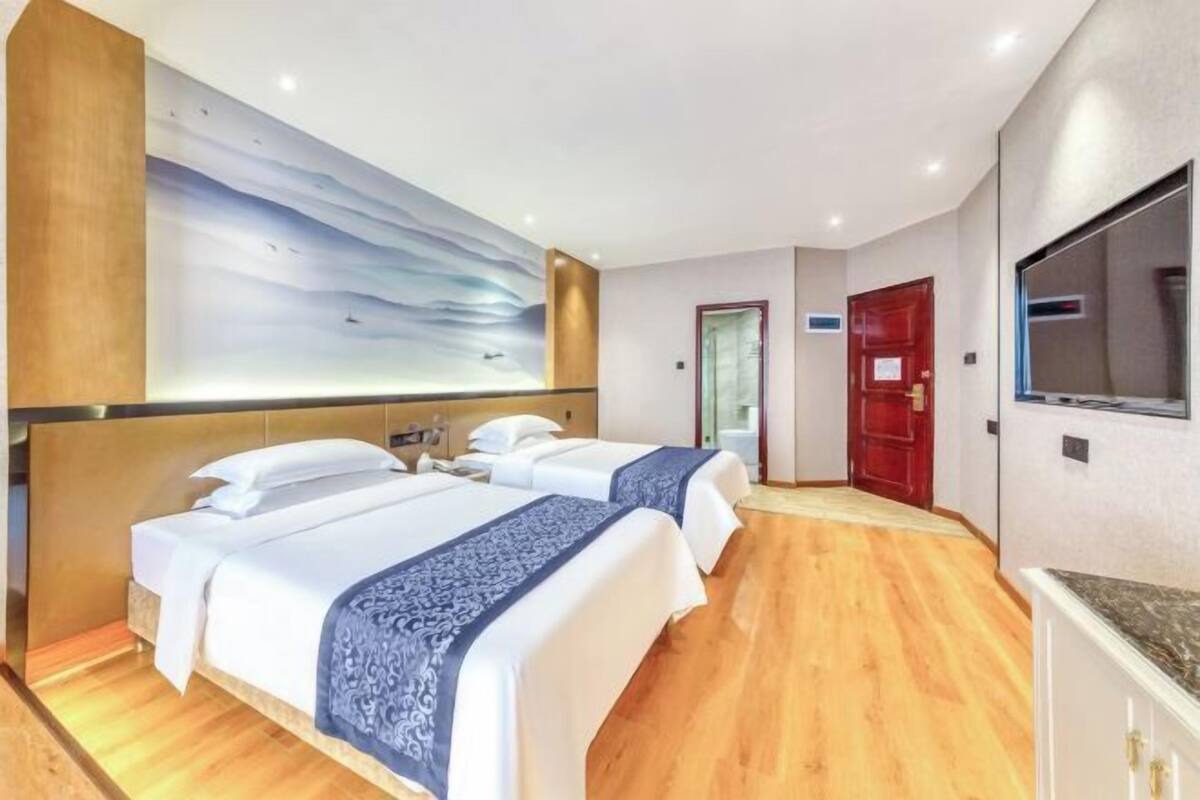 A hotel room with two beds, a large wall mural, wooden flooring, and a red door.