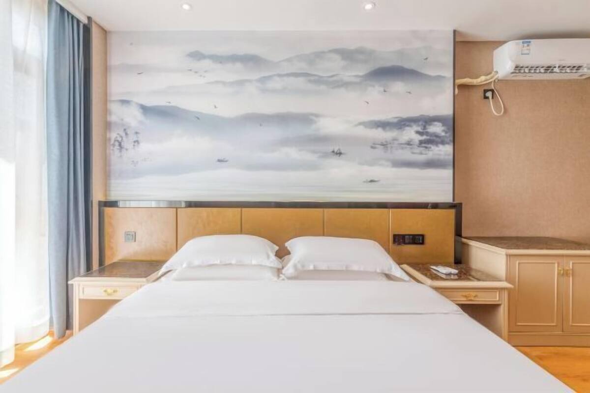 A hotel room with a bed, nightstands, a wall-mounted air conditioner, and a large painting of a mountainous landscape.