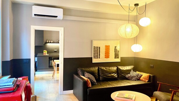 Una sala de estar moderna con un sofá de cuero negro, una mesita redonda y una estantería con libros. Se ve una cocina a través de un vano abierto.