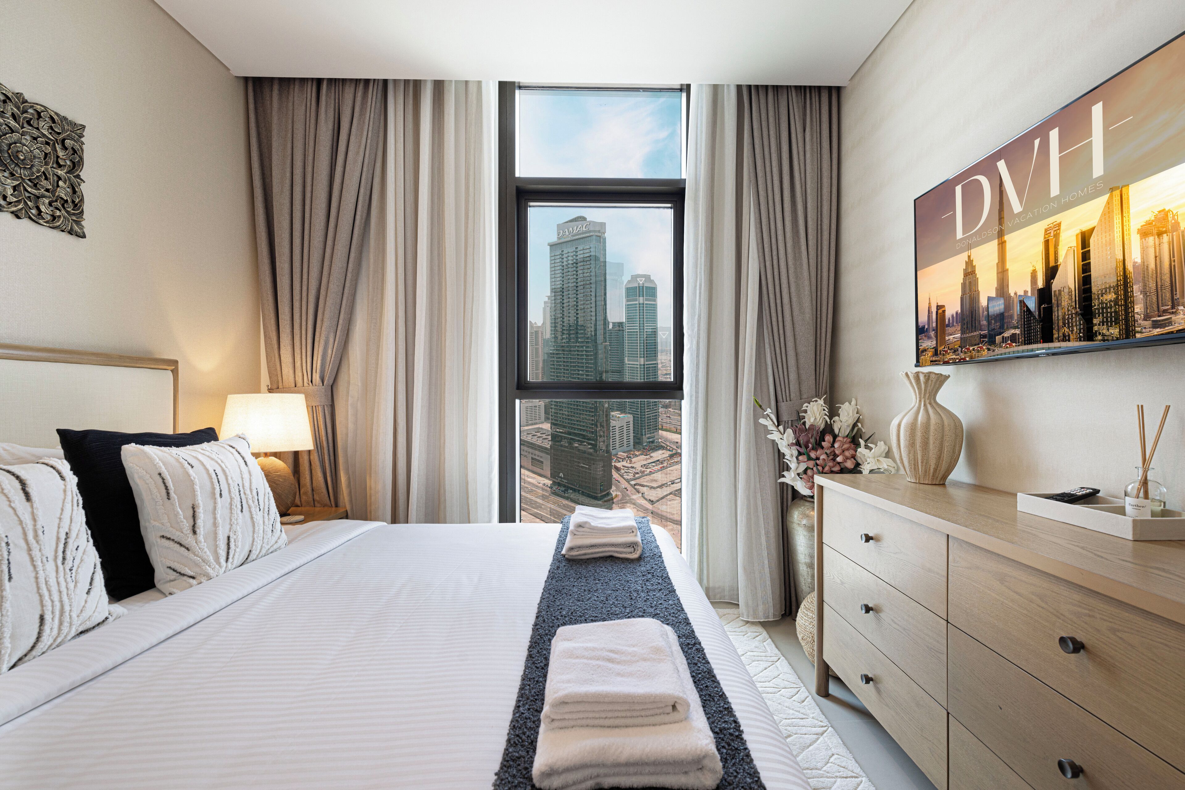 A hotel room with a large bed, a dresser, a window with a city view, and a framed picture on the wall.