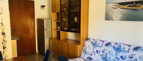 A cozy room with a wooden cabinet, a sofa with a floral pattern, a dining table, chairs, a refrigerator, and a clock on the wall.