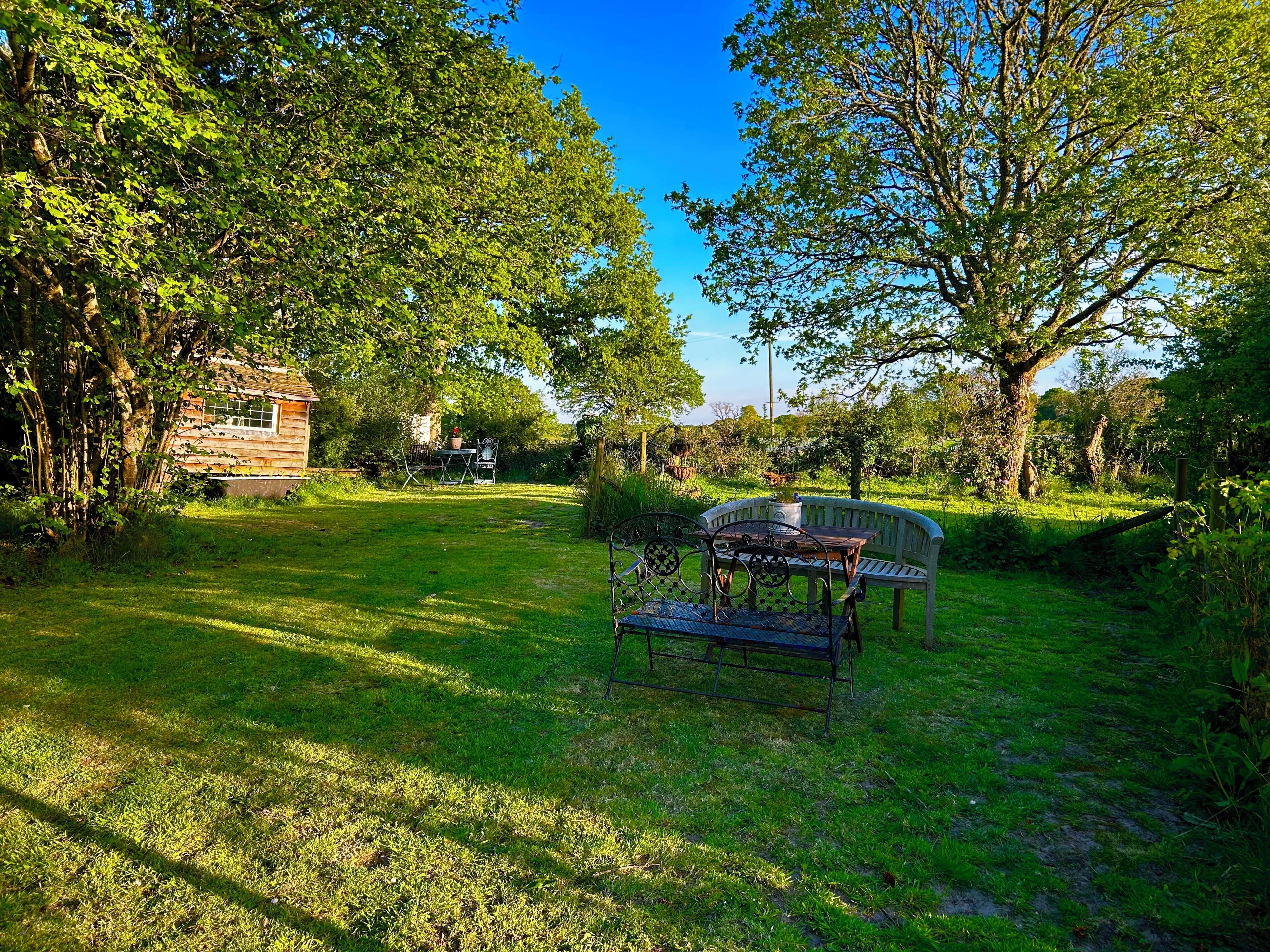 A rustic wooden cabin surrounded by lush greenery, a metal bench, and a table with chairs in a sunny outdoor setting.