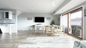 A modern kitchen with white cabinetry, a dining area with a round table and chairs, a flat-screen TV mounted on the wall, and a large sliding glass door leading to a balcony. - TideWatch-Relax in Brigantine w/Views,Beach and Pool (Brigantine)