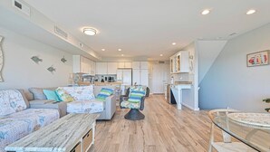 A modern living room with a sofa, armchair, and glass coffee table. A kitchen is visible in the background with white cabinets and a dining area. - TideWatch-Relax in Brigantine w/Views,Beach and Pool (Brigantine)