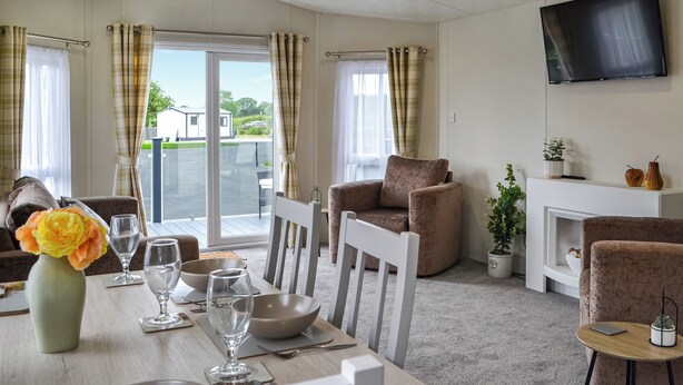 A modern living room with a dining table set for four, a comfortable armchair, a sofa, a flat-screen TV mounted on the wall, and a balcony with a view of the outside. - 3 bedroom accommodation in Carnaby, near Bridlington (Carnaby, near Bridlington)