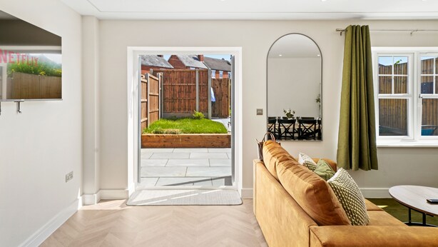 A modern living room with a sofa, a dining table, and a view of a garden through an open door. - Cove Road Houses by Redwood Stays (Farnborough)