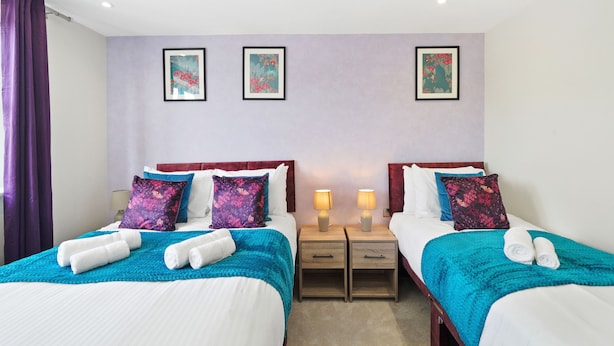 A hotel room with two beds, each with a teal bedspread and matching pillows, a nightstand with a lamp, and framed artwork on the wall. - Cove Road Houses by Redwood Stays (Farnborough)