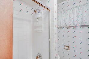 A bathroom with white tiled walls, a shower with a glass door, and a toilet with a floral-patterned curtain. - Sullivan Lake Cabin w/ Pierz & Hillman Access! (Pierz)