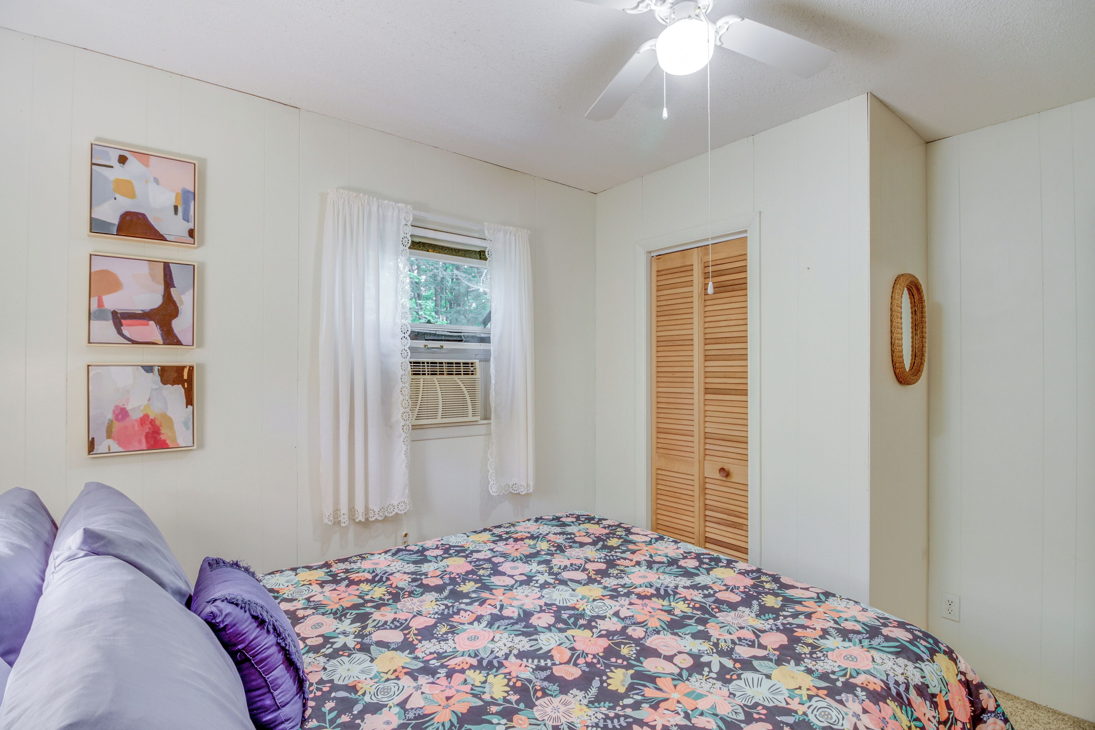 A bedroom with a floral bedspread, a ceiling fan, a window with curtains, and a wooden wardrobe.