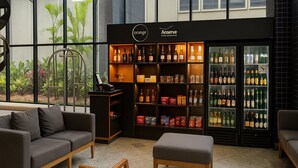 A modern lounge area with a sofa, ottoman, and chairs, a display of beverages, and a view through large windows. - Elegance Suítes Jardins (São Paulo)