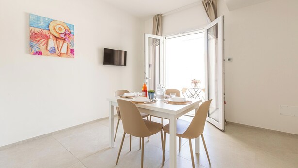 A dining area with a white table, beige chairs, a painting, and a television on the wall. - Residence Albeach - Bilo 3 (Porto Cesareo)
