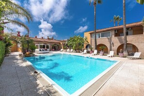A swimming pool surrounded by lounge chairs and palm trees, with a building featuring arched windows and a tiled roof in the background. - Thalissa Beach House - Mono White by BarbarHouse (Taranto)