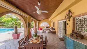 Un patio cubierto con una mesa, sillas y un ventilador de techo. Se ve una piscina a través de un arco, y una cocina con un lavamanos y armarios.