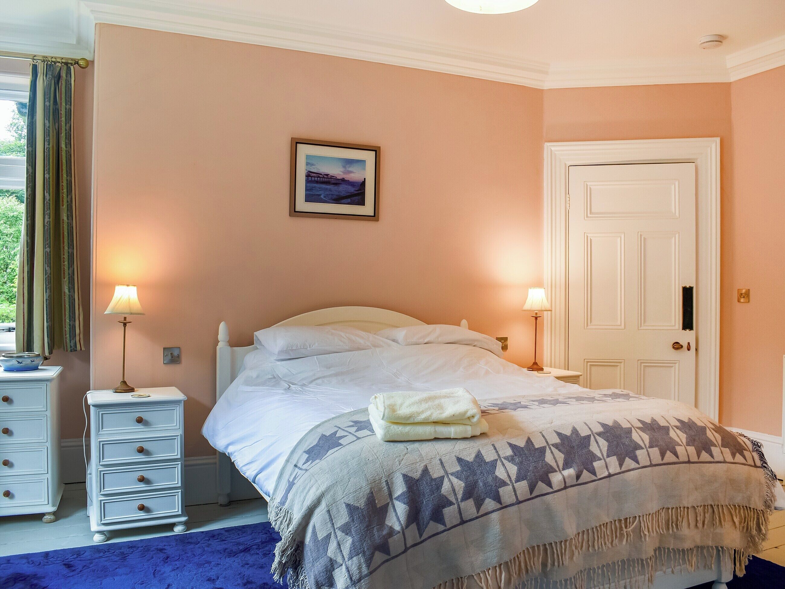 A neatly made bed with a star-patterned blanket, two bedside lamps, a framed picture on the wall, and a white door.
