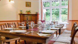 A dining room with a long wooden table set for a meal, a fireplace, and a large window with a view of greenery. - 6 bedroom accommodation in Okehampton (Okehampton)