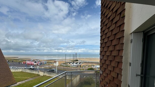A balcony with a view of a beach, a parking area, and a building with a brick facade. - Nice T2 with 2 balconies on the seafront (Ouistreham)