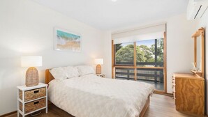 Un dormitorio con una cama grande, una mesita de noche con lámpara, un aparador de madera y una ventana con vistas a árboles.