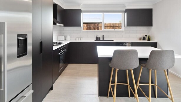 A modern kitchen with dark cabinetry, a stainless steel refrigerator, a built-in oven, and a large island with bar stools. - Fresh Seas - Mornington (Mornington)