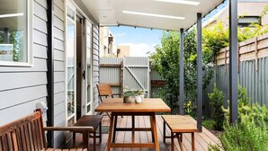 En udendørs terrasse med et træbord og stole, omgivet af et træhegn og grønne planter.