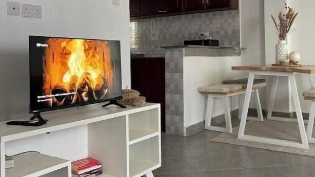 Una sala de estar moderna con un televisor de pantalla plana mostrando una chimenea, una pequeña mesa de comedor y una zona de cocina con armarios oscuros.