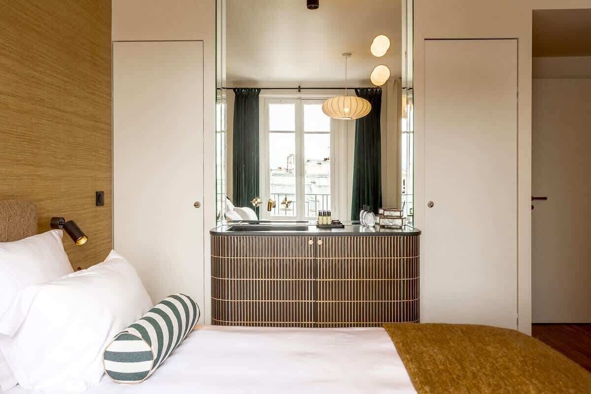 A modern hotel room with a large bed, a bedside table, a mirror, and a view of the cityscape through the window.