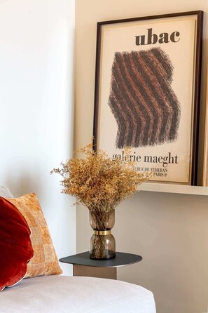 Una obra de arte en marco con la palabra “ubac” y un trazo de pincel texturizado, un jarrón con flores secas sobre una mesita y una almohada roja sobre una cama.
