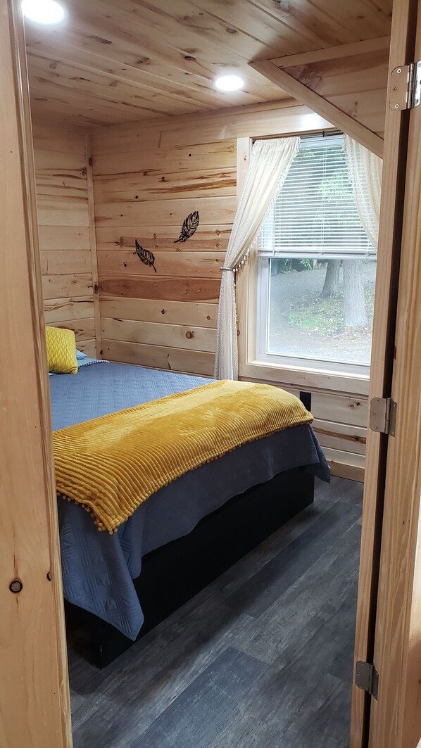 Un dormitorio de cabaña de madera con una cama, una ventana con persianas y una lámpara en el techo.