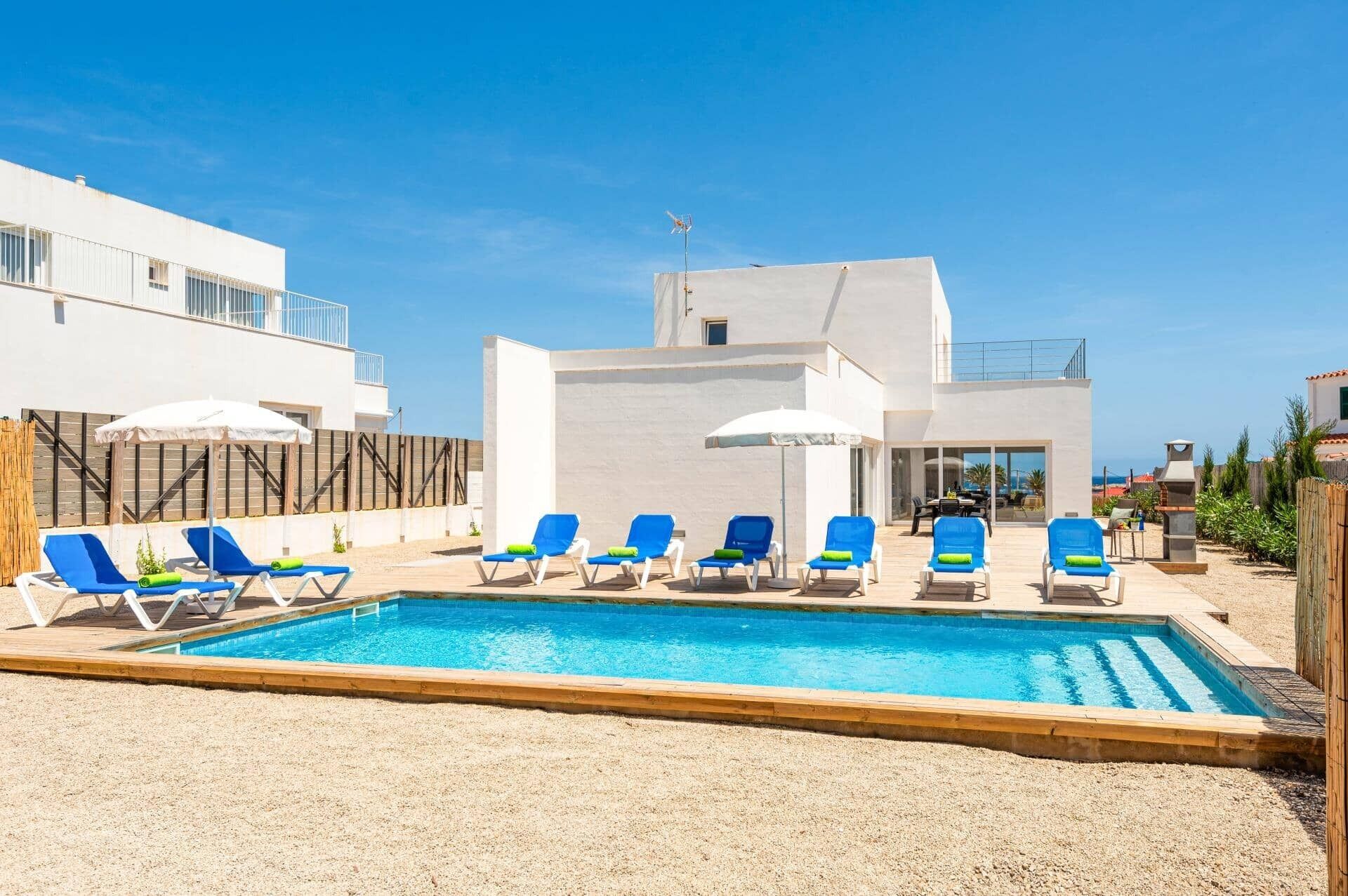 Un espace piscine moderne, avec des chaises longues bleues et des parasols, entouré d’un bâtiment blanc doté d’un toit plat et d’une antenne parabolique bien visible.