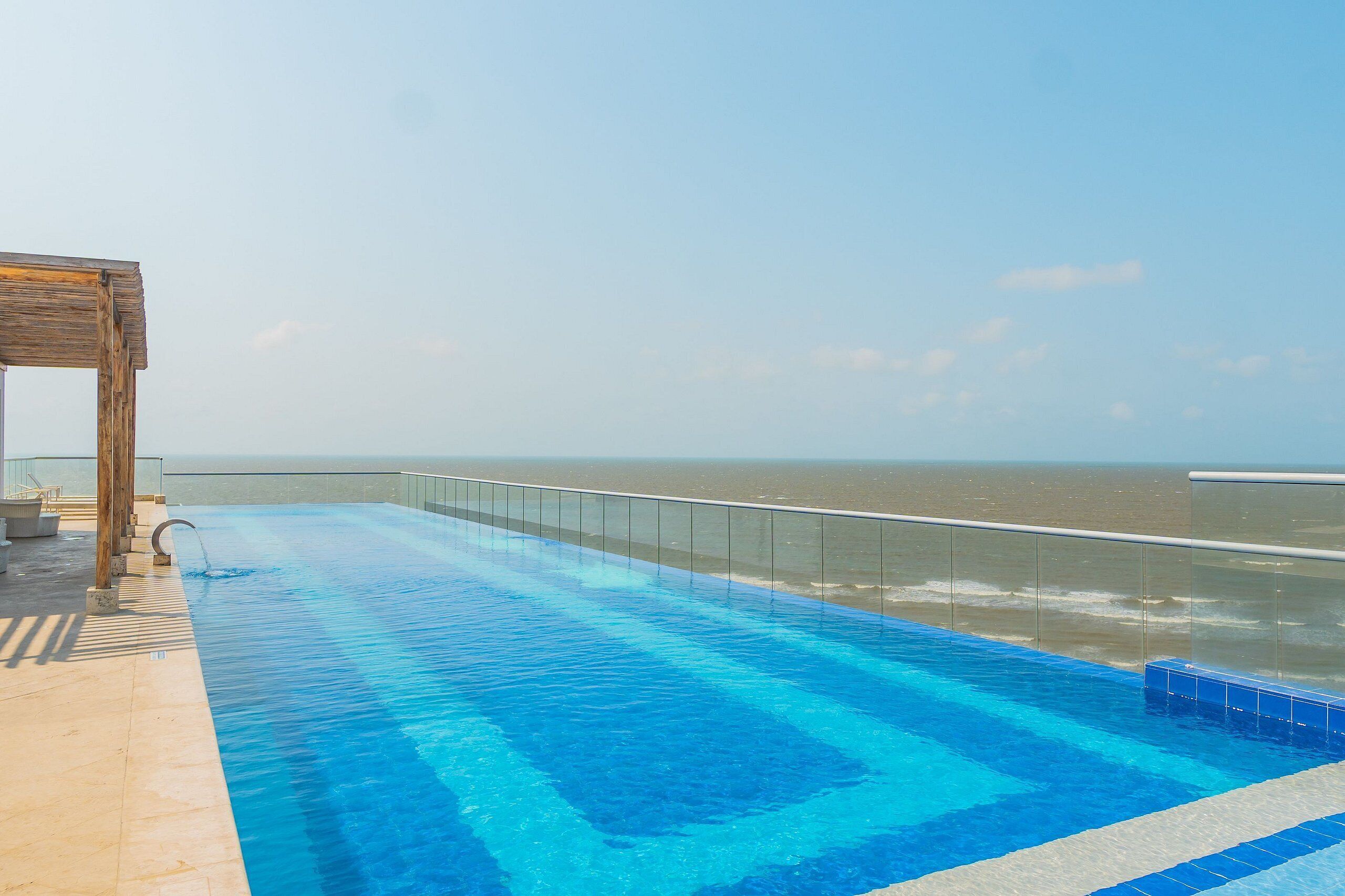 A swimming pool with a glass barrier, a wooden pergola, and a view of the sea.