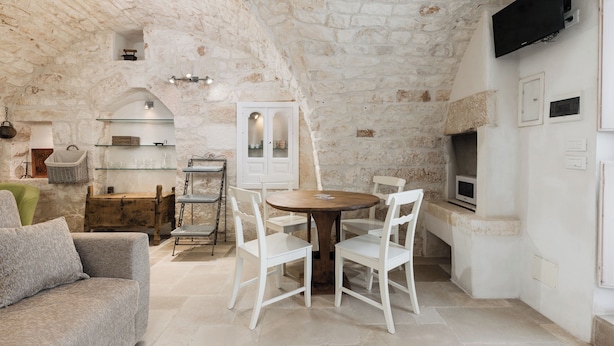 Una habitación rústica con paredes de piedra, una mesa redonda de madera y sillas blancas, un sofá verde y una pequeña cocina con microondas.