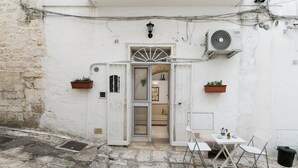Una calle estrecha con un edificio blanco que cuenta con una pequeña zona de estar al aire libre, una mesa, sillas, una planta en maceta y un equipo de aire acondicionado.