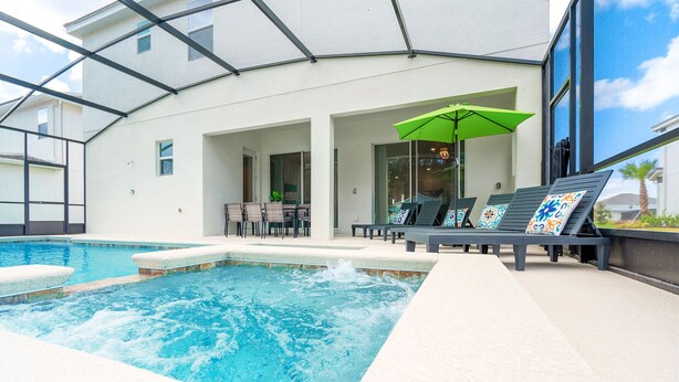 An indoor swimming pool with lounge chairs and a table, surrounded by a glass-enclosed patio area.