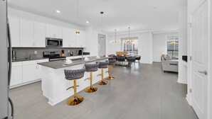 A modern kitchen with white cabinets, a central island with bar stools, and a dining area with a sofa and table. - Last Minute Disney! Private Pool, Hot Tub, Game Room & Themed Bedrooms. Book Now! #108421 (Orlando)