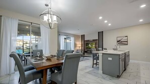 A modern kitchen with a central island, a dining area with a wooden table and grey chairs, and a view of the outdoors through large windows. - Last Minute Disney! Townhouse with Private Pool. Book Now! #CG3435 (Orlando)