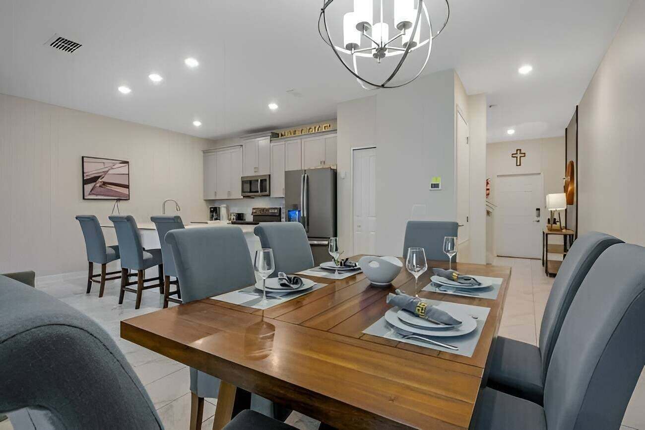A modern dining room with a wooden table set for six, surrounded by grey chairs. A kitchen with white cabinets and a microwave is visible in the background.