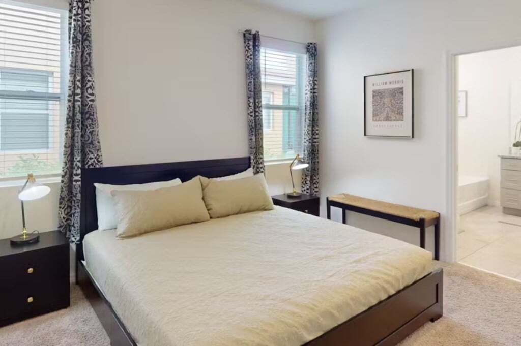 Un dormitorio con cama, dos mesitas de noche, una lámpara de noche, una ventana con cortinas y un cuadro en la pared.