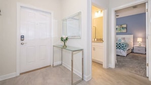 Un pasillo con una puerta blanca, un tocador de cristal con un jarrón de flores, y un dormitorio con cama y mesita de noche.