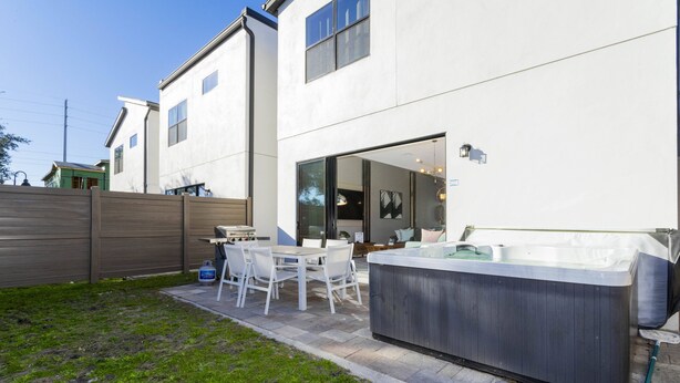 A modern outdoor patio with a hot tub, a dining area with chairs, and a large window overlooking the yard. - Disney! Private Pool, Hot Tub & Themed Bedrooms. Book Now! #VDR14 (Orlando)