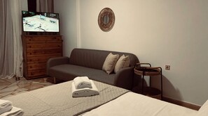 A hotel room with a bed, a grey sofa, a wooden dresser, a television, and a wall-mounted mirror.