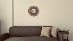 A grey sofa with two striped pillows against a plain wall, a woven mirror above it, and a wicker side table with a lamp. - Apartment in the Heart of Tinos Town for 4 Guests (Tinos)
