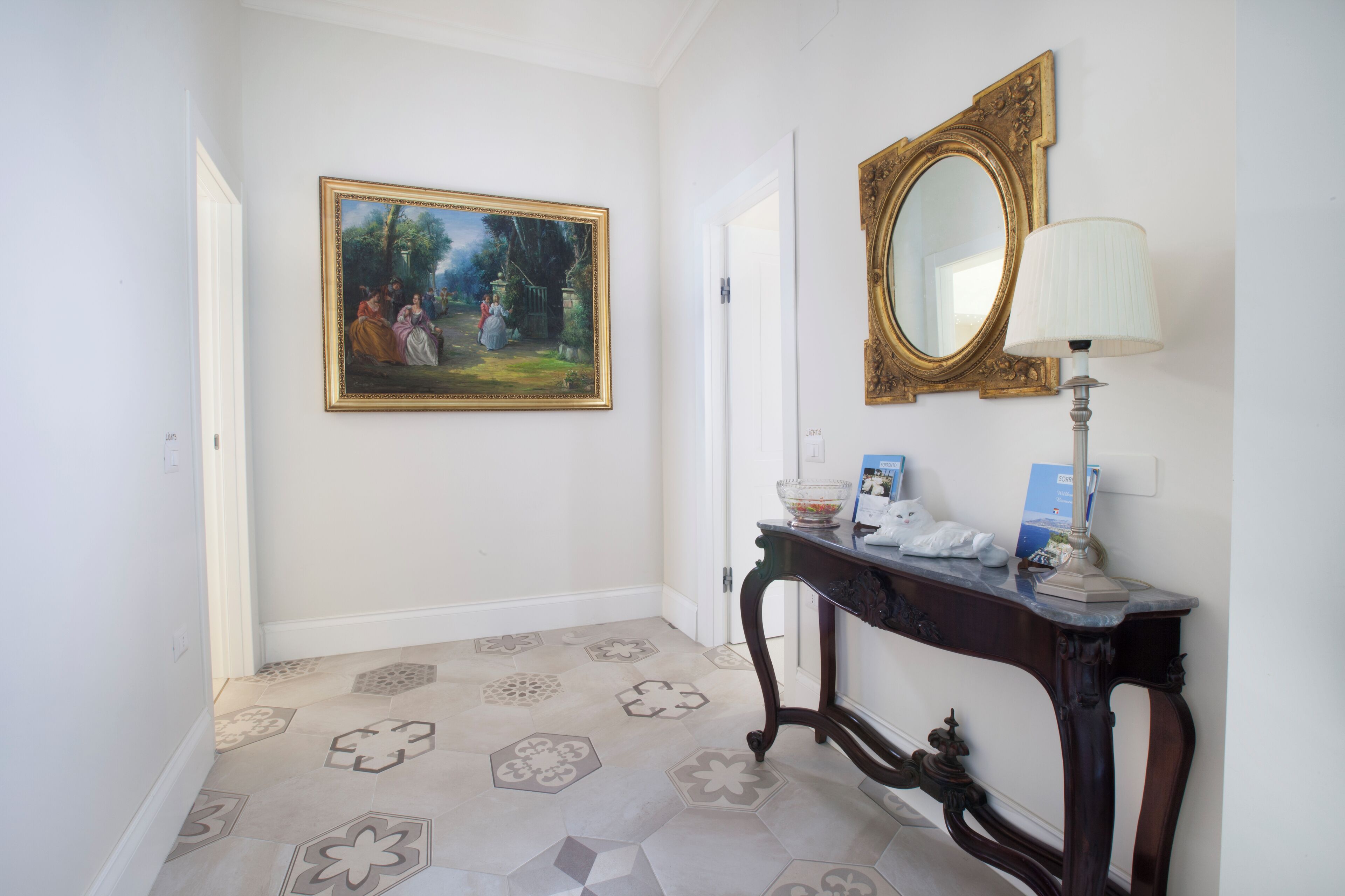 A hallway with a tiled floor, a framed painting, a mirror, and a console table with a lamp and a cat figurine.