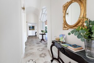 Un pasillo con suelo estampado, un espejo, una mesita consola con libros y un florero, y una puerta que da a otra habitación.