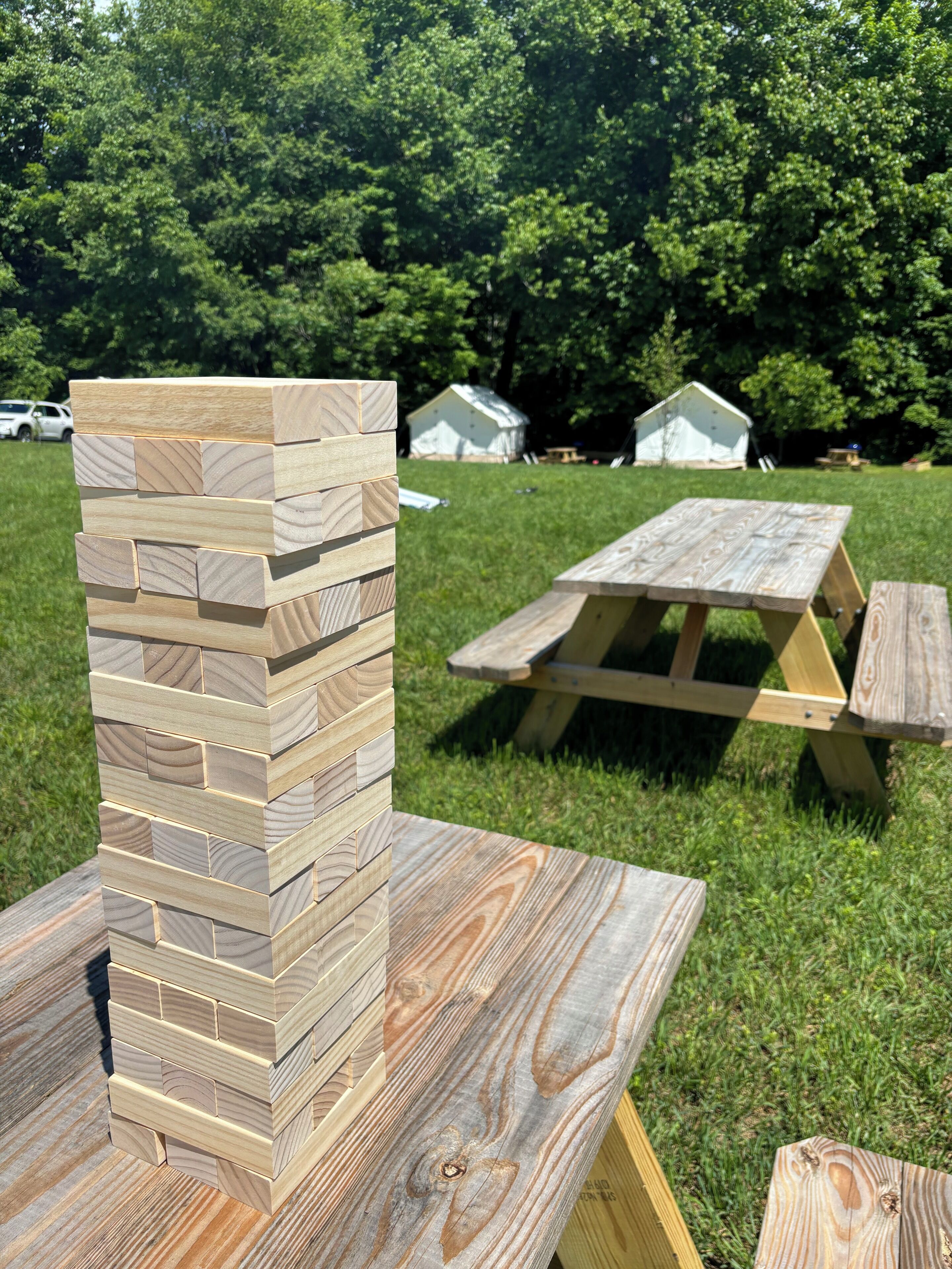 A stack of wooden blocks and a picnic table in a grassy outdoor area with trees in the background.