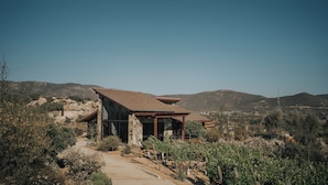 A modern house with a stone facade, large windows, and a sloped roof, surrounded by a dry landscape and hills. - Cabins by 7 Valles (Valle de Guadalupe)