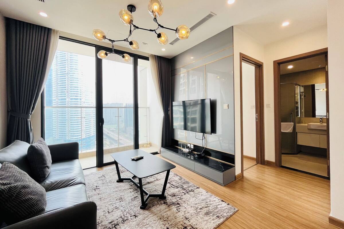 Un salon moderne avec un canapé, une table basse et un téléviseur. On peut voir une grande fenêtre avec des rideaux, un balcon et une salle de bain.