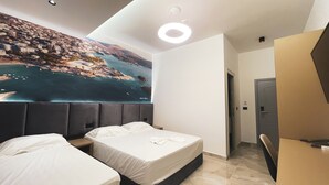 A hotel room with two beds, a large mural of a coastal town, a modern ceiling light, and a door to the right. - Hotel Ruko Boutique (Golem)