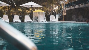 A swimming pool with white lounge chairs and umbrellas, surrounded by trees and a decorative wall. - Hotel Ruko Boutique (Golem)